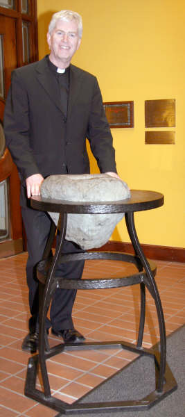 The Very Rev Colm  McBride pictured at the original Holy Water Font found during excavations of the site circa 1850.  The brass plaques on the wall behind Fr. Hickland Fr McBride gives details of the history of the font.